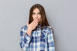 Portrait of a young woman covering her mouth with palm and looking away over gray background